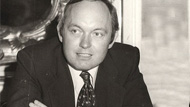 black and white photo of clean shaven dark haired man in a suit sitting down with arms crossed 