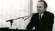 black and white photo of a dark haired man in a suit with a tie infront of a microphone 